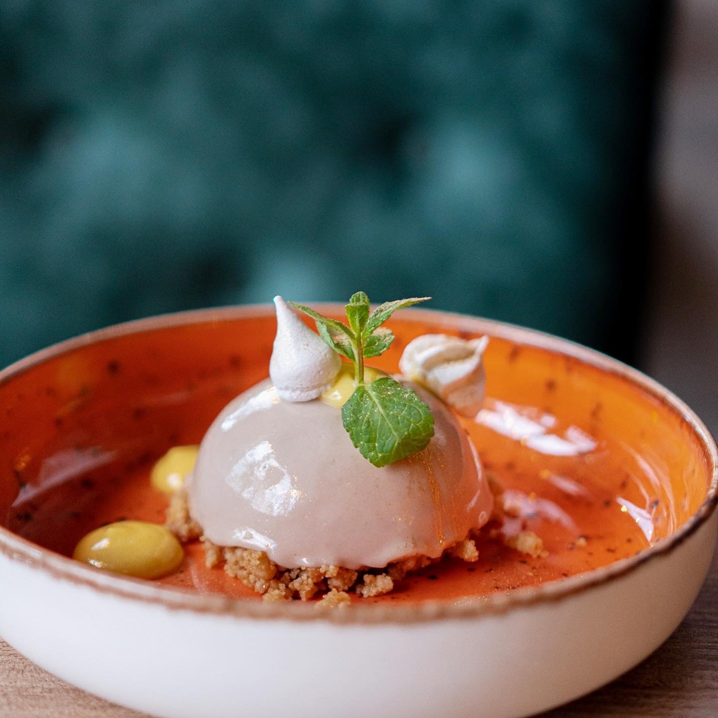 Elegancko podany deser: czekoladowa kula na kruchym spodzie, udekorowana mini bezami, listkiem mięty i kroplami żółtego kremu, serwowana w pomarańczowo-białej miseczce na drewnianym stole,...