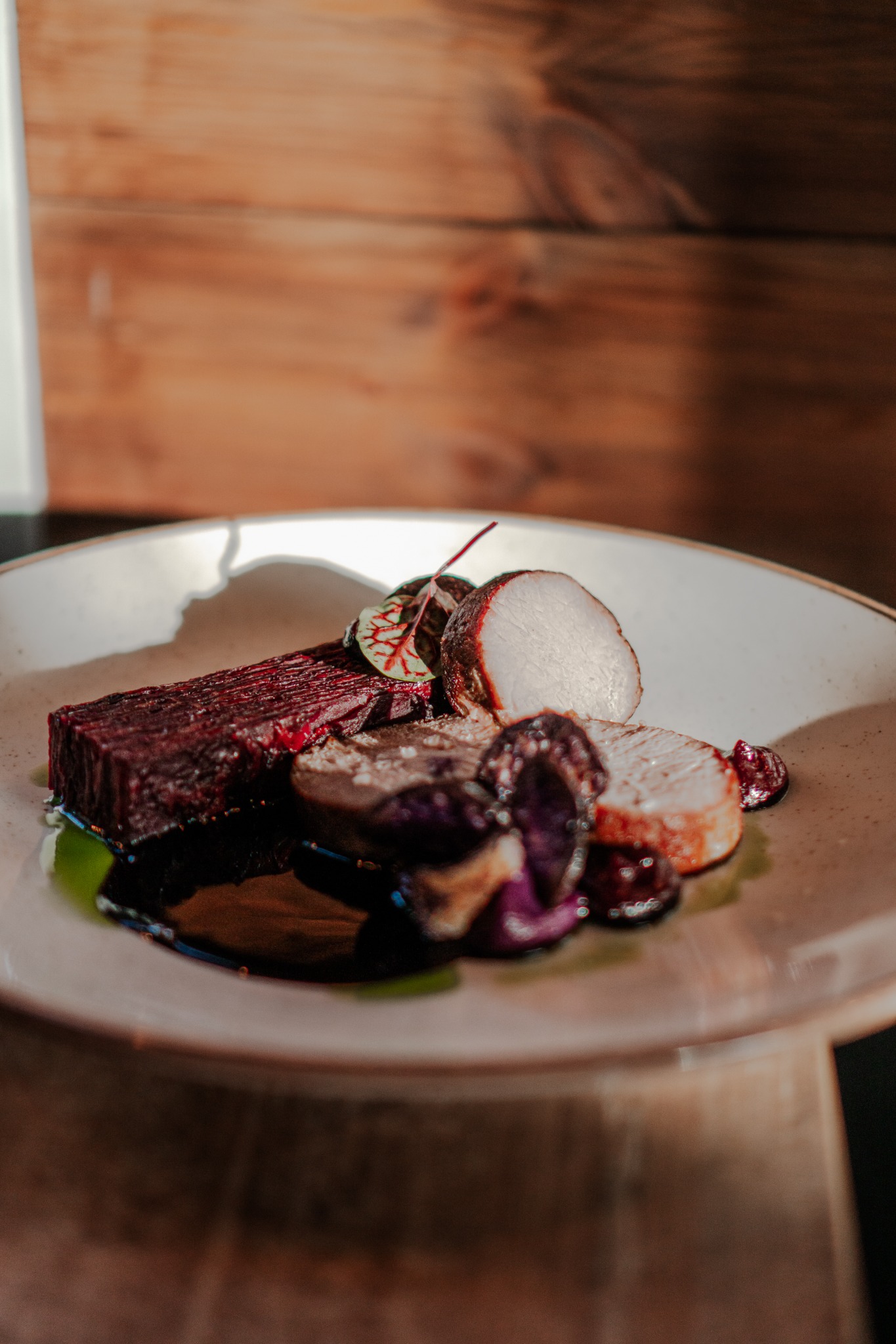 Elegancko podane danie na talerzu: plasterki pieczonego buraka, purpurowe warzywa i jasne mięso polane ciemnym sosem na tle drewnianej ściany.