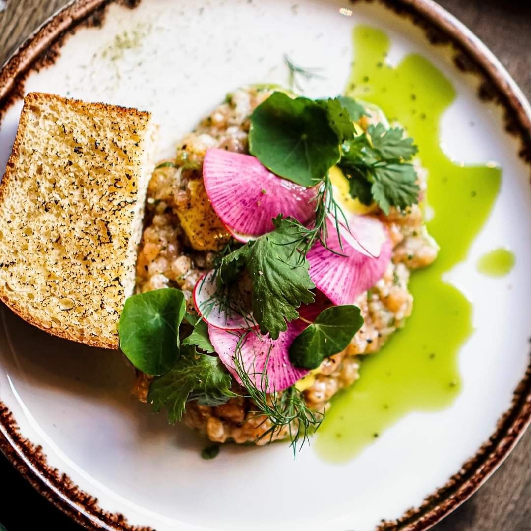 Elegancko podany tatar z łososia z rzodkiewką, ziołami i sosem pistacjowym obok grzanki na talerzu z brązową obwódką.