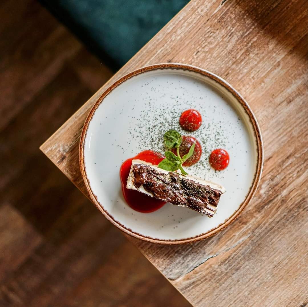 Elegancko podany deser na białym talerzu z brązową obwódką: prostokątny kawałek ciasta polany czekoladą, otoczony sosem malinowym, trzema okrągłymi kroplami sosu, posypany zielonym proszkiem...