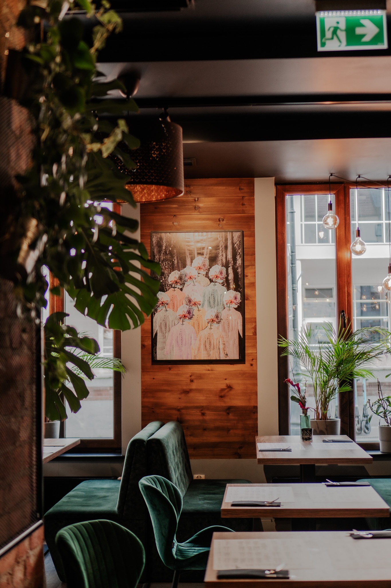 Wnętrze restauracji z drewnianymi panelami, zielonymi aksamitnymi meblami, roślinami doniczkowymi i abstrakcyjnym obrazem na ścianie.