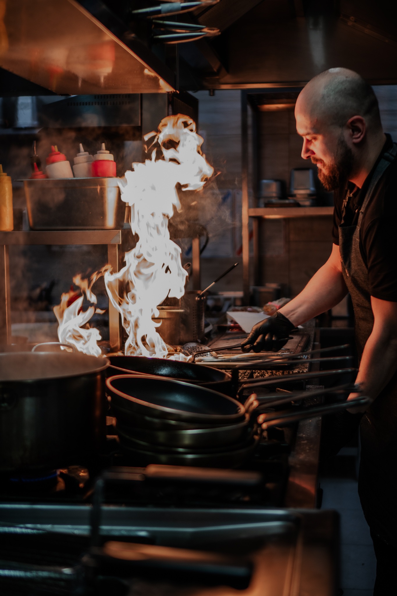 Kucharz w czarnych rękawicach flambirowuje potrawę na palniku w profesjonalnej kuchni, obok stos patelni.