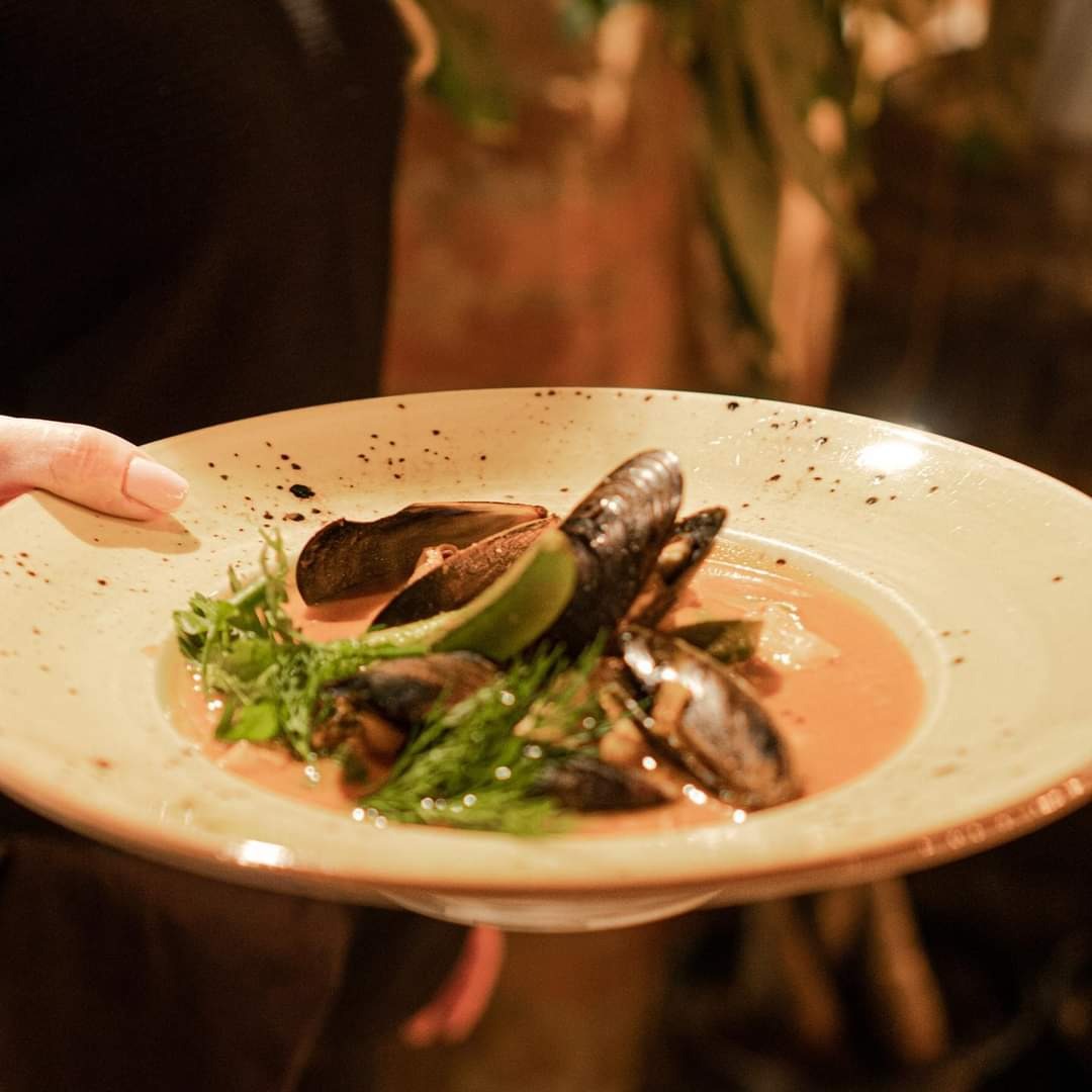 Kelnerka trzyma talerz z małżami w pomidorowym sosie z limonką i koperkiem, na talerzu w stylu rustykalnym z ciemnymi plamkami.