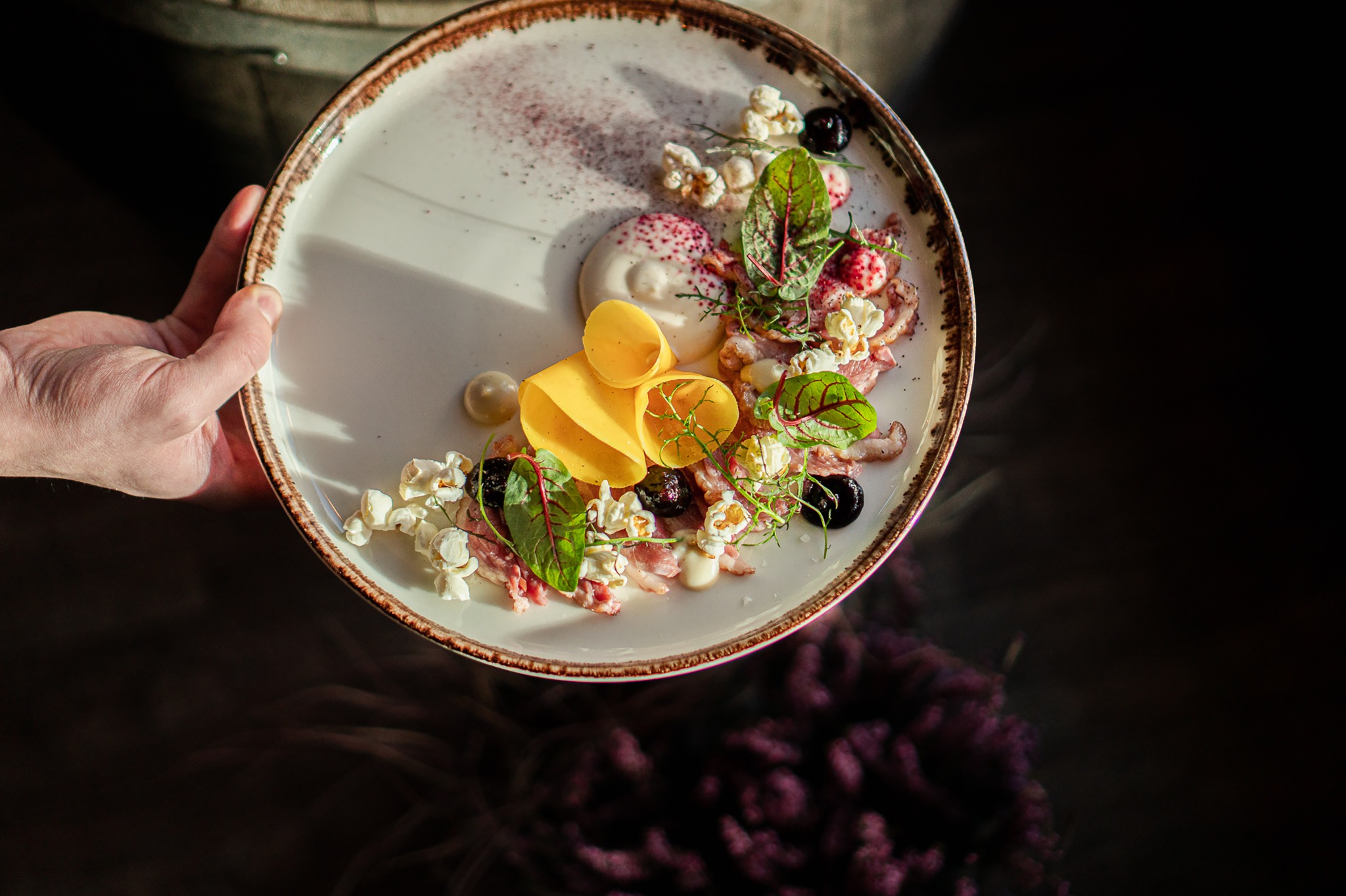Elegancko podane danie na talerzu trzymanym przez kelnera: cienkie plastry mięsa, krem, popcorn, czarne oliwki i listki ziół, udekorowane pudrem, na tle ciemnego wnętrza.