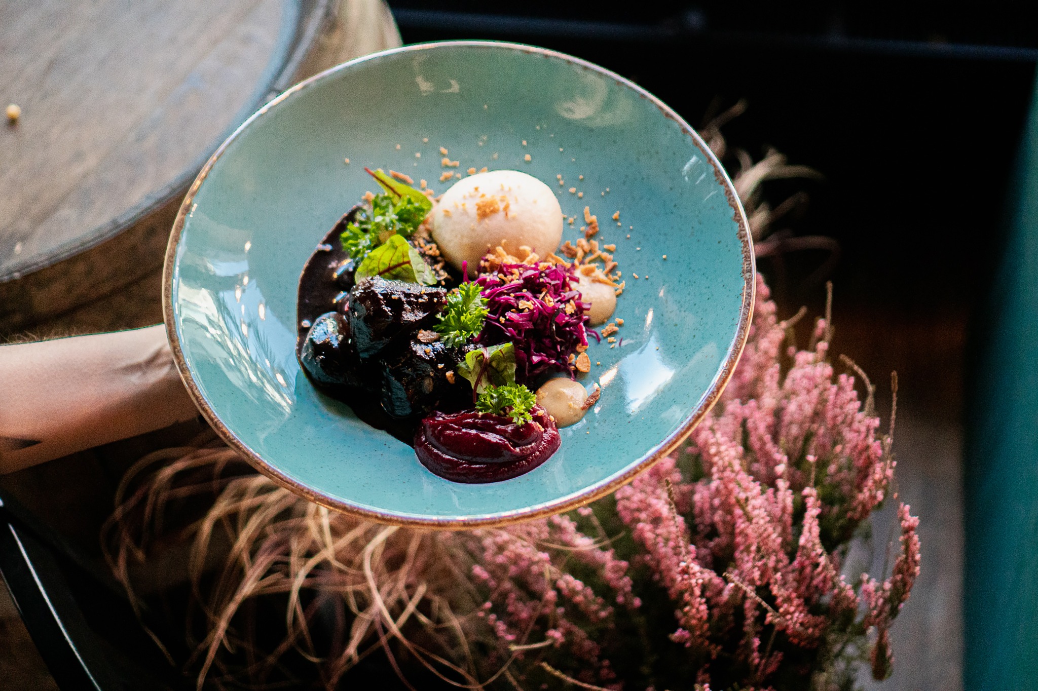 Elegancko podane danie mięsne z ciemnym sosem, puree, czerwoną kapustą i dekoracją z ziół na turkusowym talerzu, trzymane w dłoni, z wrzosem w tle.