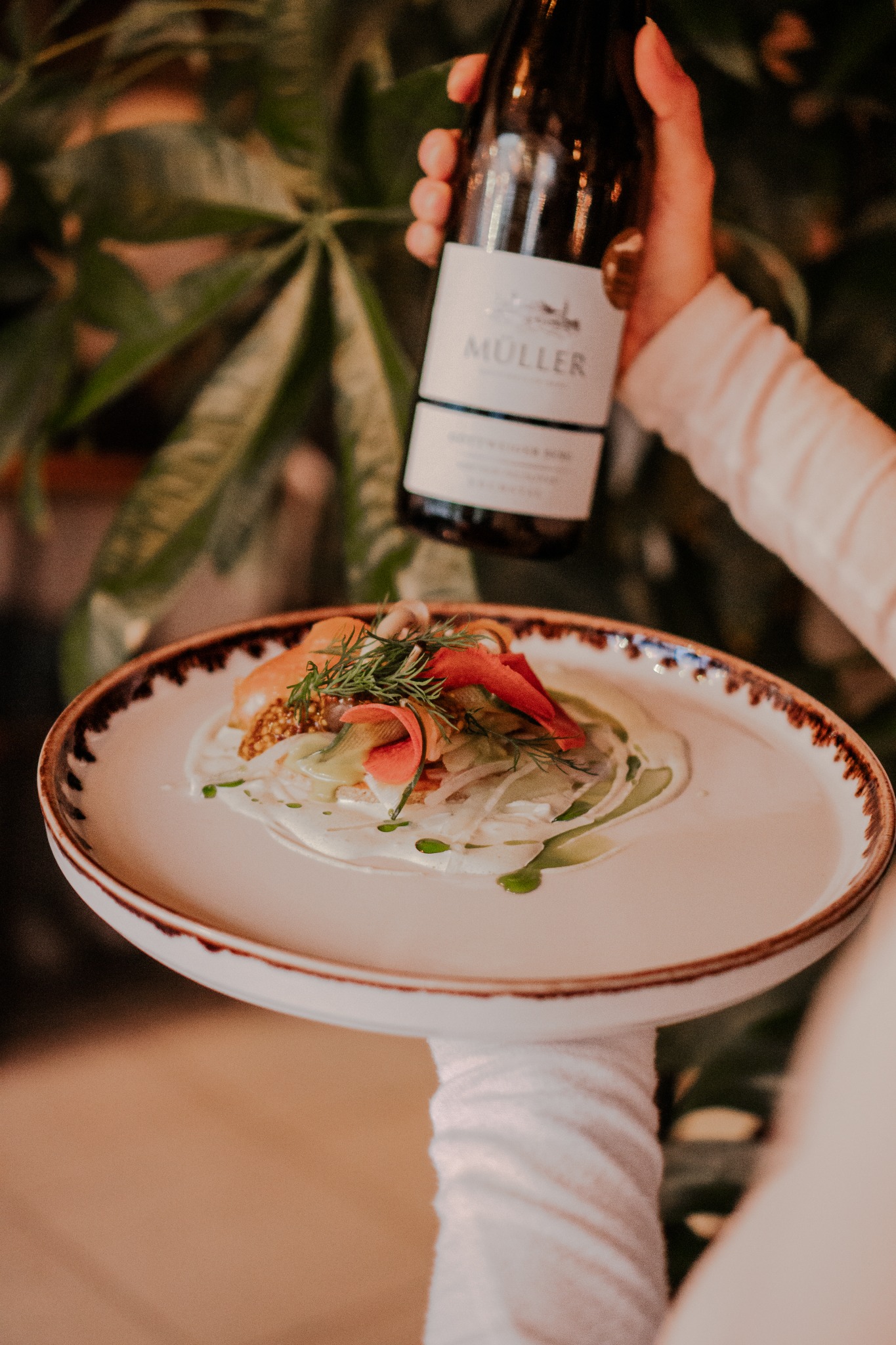 Elegancko podane danie z rybą i warzywami na białym talerzu z ciemnym brzegiem, trzymane przez kelnera w białej rękawiczce, w tle butelka wina marki Muller, serwowane w restauracji w Sopocie.