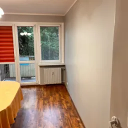 Pomalowane na szaro wnętrze pokoju z parkietem, widok na okno balkonowe z czerwonymi roletami i grzejnik.