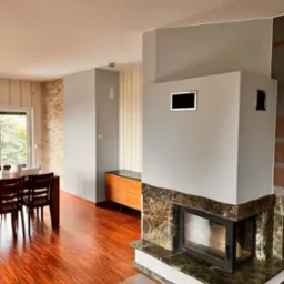 Jasne wnętrze po metamorfozie: ściany w odcieniach szarości i beżu, drewniana podłoga, marmurowy kominek z szybą, stół z krzesłami przy oknie, szafka z drewnianymi frontami.