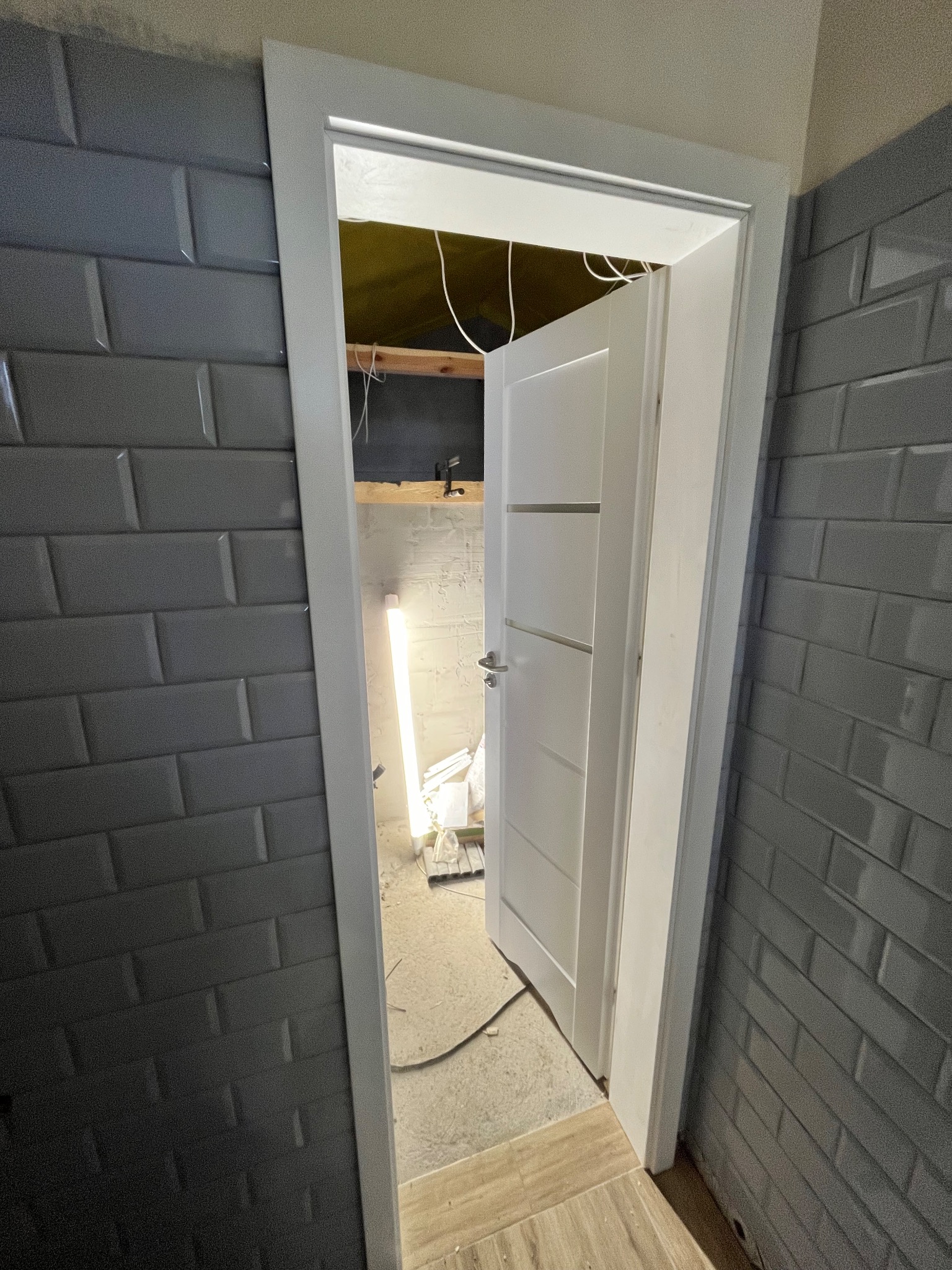 Otwarta, biała, nowoczesna drzwi wewnętrzne zamontowane w ścianie wyłożonej szarą, cegiełkową glazurą w trakcie prac wykończeniowych, widoczne kable i tymczasowe oświetlenie.