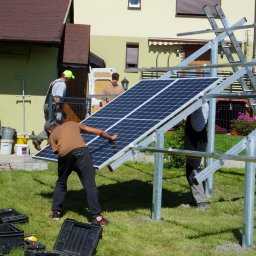 ODNAWIALNE ŹRÓDŁA ENERGII SPÓŁKA Z OGRANICZONĄ ODPOWIEDZIALNOŚCIĄ - Montaż paneli słonecznych na metalowej konstrukcji w przydomowym ogrodzie, widoczni trzej instalatorzy w trakcie pracy, skrzynki z narzędziami na trawie.