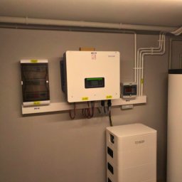 ODNAWIALNE ŹRÓDŁA ENERGII SPÓŁKA Z OGRANICZONĄ ODPOWIEDZIALNOŚCIĄ - Instalacja domowa: biały inwerter Sofar i magazyn energii Dyness na szarej ścianie, widoczna skrzynka elektryczna i rury instalacyjne pod sufitem.