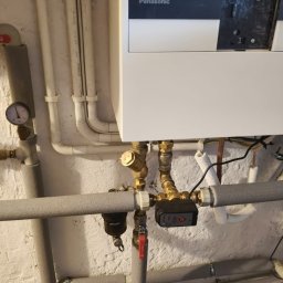 ODNAWIALNE ŹRÓDŁA ENERGII SPÓŁKA Z OGRANICZONĄ ODPOWIEDZIALNOŚCIĄ - Biały piec Panasonic z podłączeniami hydraulicznymi i rurami w szarej izolacji na tle białej, chropowatej ściany. Widoczny manometr i zawór.