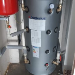 ODNAWIALNE ŹRÓDŁA ENERGII SPÓŁKA Z OGRANICZONĄ ODPOWIEDZIALNOŚCIĄ - Szary, cylindryczny zbiornik Cordivari z manometrem i licznymi przyłączami, obok czerwony zbiornik wyrównawczy, całość w pomieszczeniu technicznym.