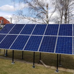 ODNAWIALNE ŹRÓDŁA ENERGII SPÓŁKA Z OGRANICZONĄ ODPOWIEDZIALNOŚCIĄ - Instalacja paneli fotowoltaicznych na konstrukcji naziemnej w ogrodzie. Widoczny dom z czerwoną dachówką i drabina. Częściowo widoczna osoba montująca panele.