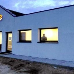 ODNAWIALNE ŹRÓDŁA ENERGII SPÓŁKA Z OGRANICZONĄ ODPOWIEDZIALNOŚCIĄ - Biały budynek z logo firmy na dachu, widoczne okna z osobą w środku i zamontowany klimatyzator na ścianie bocznej. Ujęcie z zewnątrz, wieczorne światło.