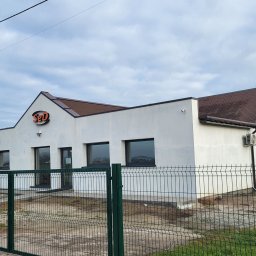 ODNAWIALNE ŹRÓDŁA ENERGII SPÓŁKA Z OGRANICZONĄ ODPOWIEDZIALNOŚCIĄ - Biały budynek z brązowym dachem i logo 'SeD' nad wejściem, otoczony zielonym ogrodzeniem. Widoczny klimatyzator na ścianie i panele słoneczne w tle.