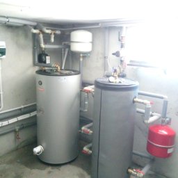 ODNAWIALNE ŹRÓDŁA ENERGII SPÓŁKA Z OGRANICZONĄ ODPOWIEDZIALNOŚCIĄ - Dwa zbiorniki buforowe z instalacją hydrauliczną w kotłowni. Widoczne rury, zawory i osprzęt. Jeden zbiornik srebrny, drugi szary z czerwonym naczyniem wzbiorczym.