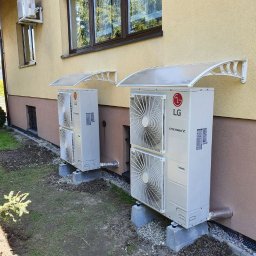 ODNAWIALNE ŹRÓDŁA ENERGII SPÓŁKA Z OGRANICZONĄ ODPOWIEDZIALNOŚCIĄ - Dwie białe pompy ciepła LG Thermav V zamontowane na betonowych podstawach przy ścianie budynku z żółtą elewacją, osłonięte przezroczystymi daszkami.