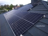 Panel fotowoltaiczny na dachu z blachodachówki, widoczny komin i drzewa w tle.