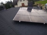 Panele słoneczne zamontowane na dachu pokrytym gontem bitumicznym, widoczne elementy montażowe i odbicia w szybach paneli, w tle dom jednorodzinny i drzewa.