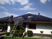 Czarny panel słoneczny na dachu domu jednorodzinnego z drabina opartą o dach, widoczne krzewy i błękitne niebo z chmurami.