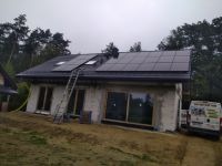 Montaż paneli słonecznych na dachu domu jednorodzinnego. Widoczna drabina oparta o dach i częściowo o elewację budynku. W tle drzewa i pochmurne niebo. Na pierwszym planie teren budowy.