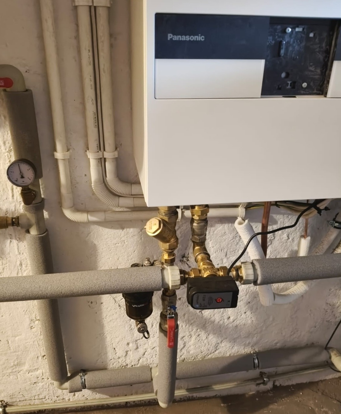 Biały piec Panasonic z podłączeniami hydraulicznymi i rurami w szarej izolacji na tle białej, chropowatej ściany. Widoczny manometr i zawór.