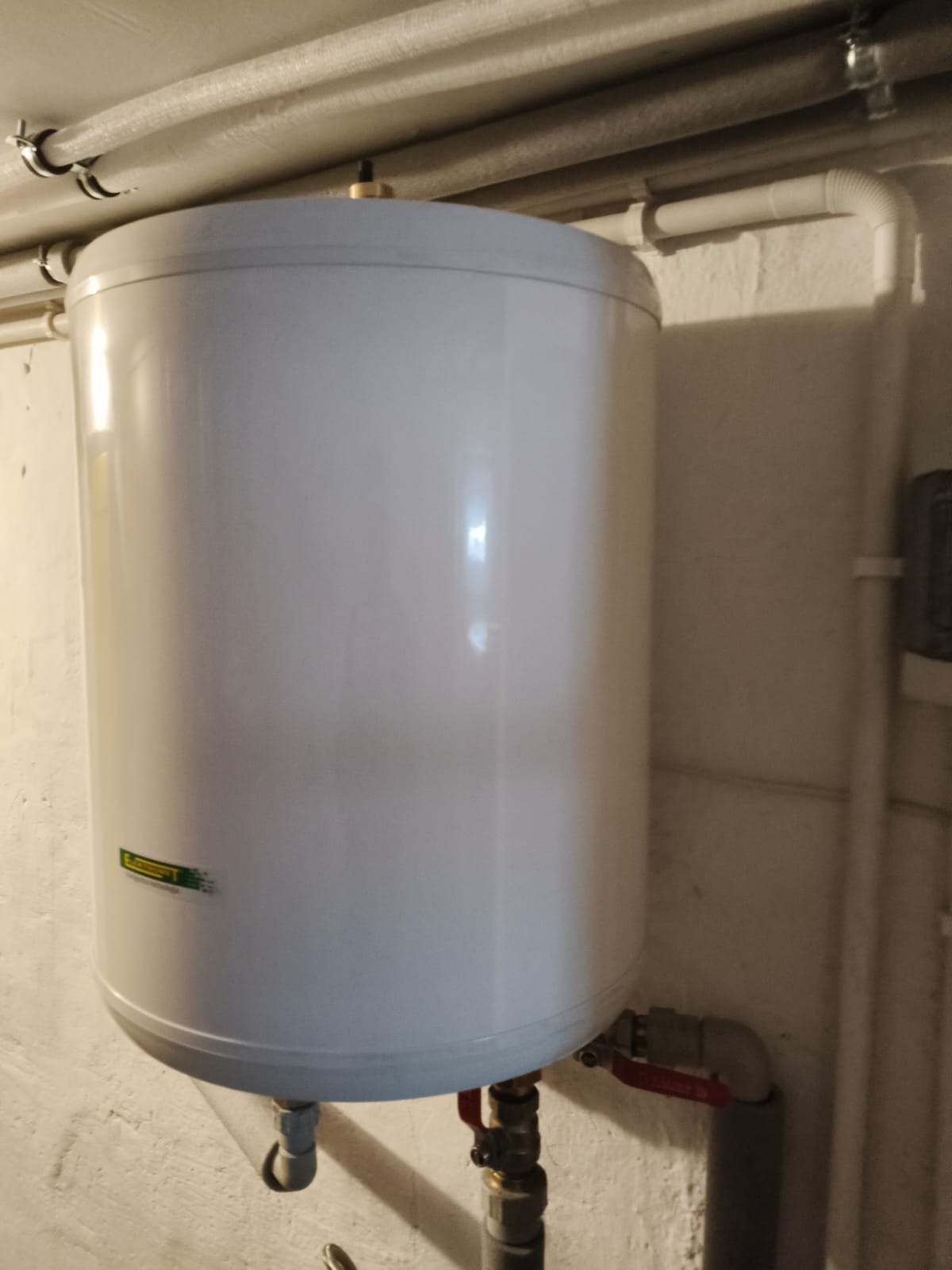 Biały, cylindryczny bojler elektryczny z widocznymi rurami doprowadzającymi wodę, zamontowany na ścianie w pomieszczeniu technicznym. Widoczne przyłącza i zawory.