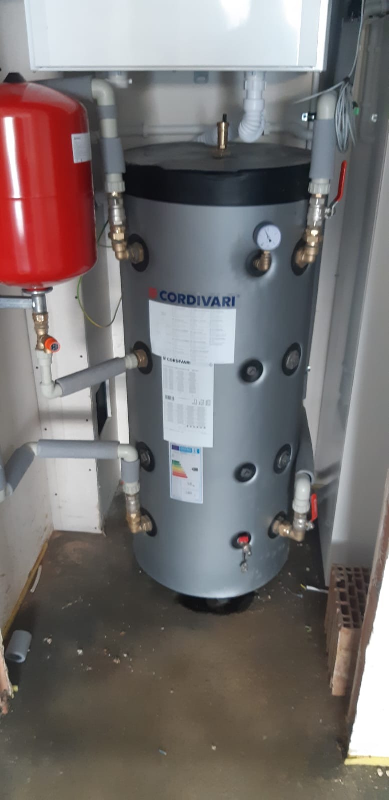 Szary, cylindryczny zbiornik Cordivari z manometrem i licznymi przyłączami, obok czerwony zbiornik wyrównawczy, całość w pomieszczeniu technicznym.