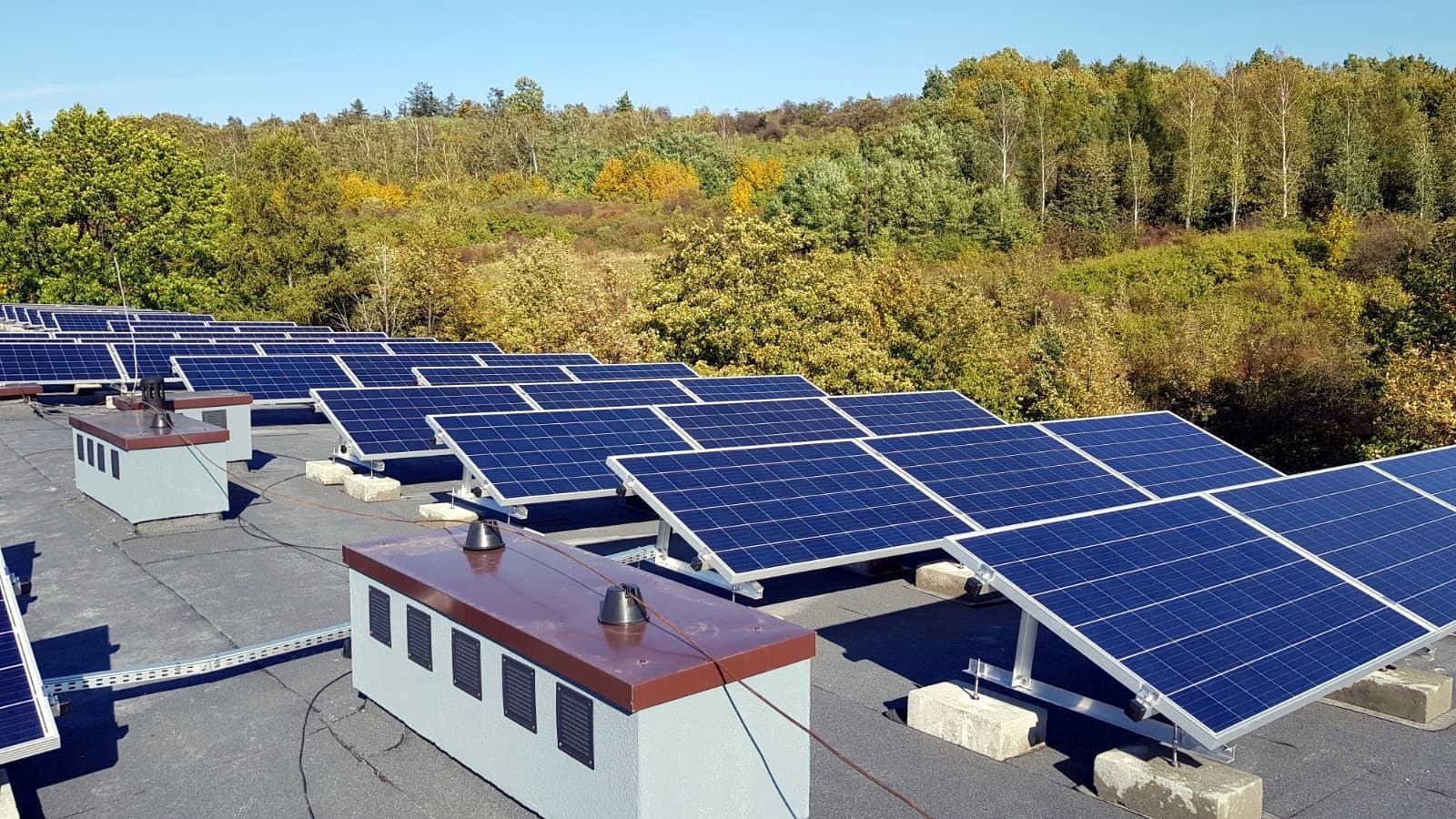 Instalacja fotowoltaiczna na dachu budynku z widocznymi panelami słonecznymi, konstrukcją wsporczą i elementami infrastruktury elektrycznej. W tle drzewa i błękitne niebo.