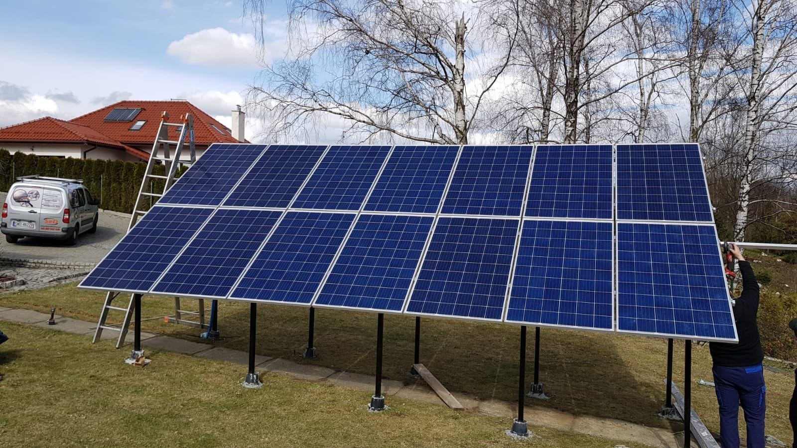Instalacja paneli fotowoltaicznych na konstrukcji naziemnej w ogrodzie. Widoczny dom z czerwoną dachówką i drabina. Częściowo widoczna osoba montująca panele.