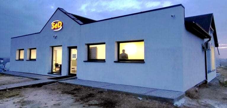 Biały budynek z logo firmy na dachu, widoczne okna z osobą w środku i zamontowany klimatyzator na ścianie bocznej. Ujęcie z zewnątrz, wieczorne światło.