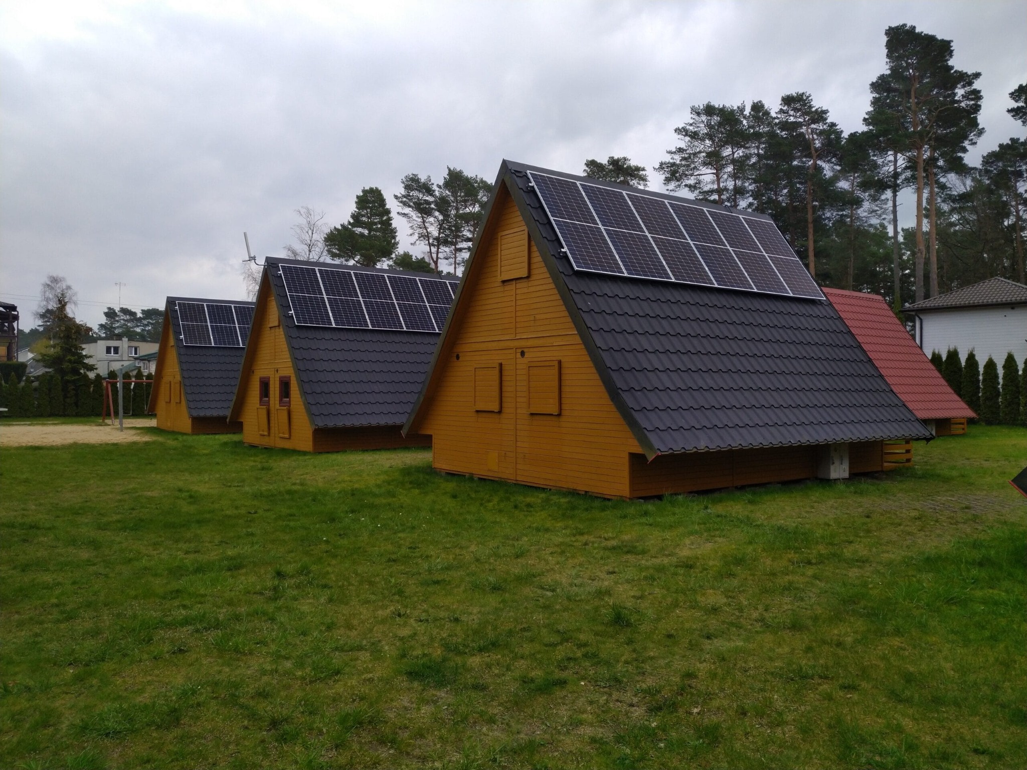 Rząd identycznych, drewnianych domków letniskowych z panelami słonecznymi na dachach, otoczonych zielonym trawnikiem i drzewami w tle, pochmurny dzień.