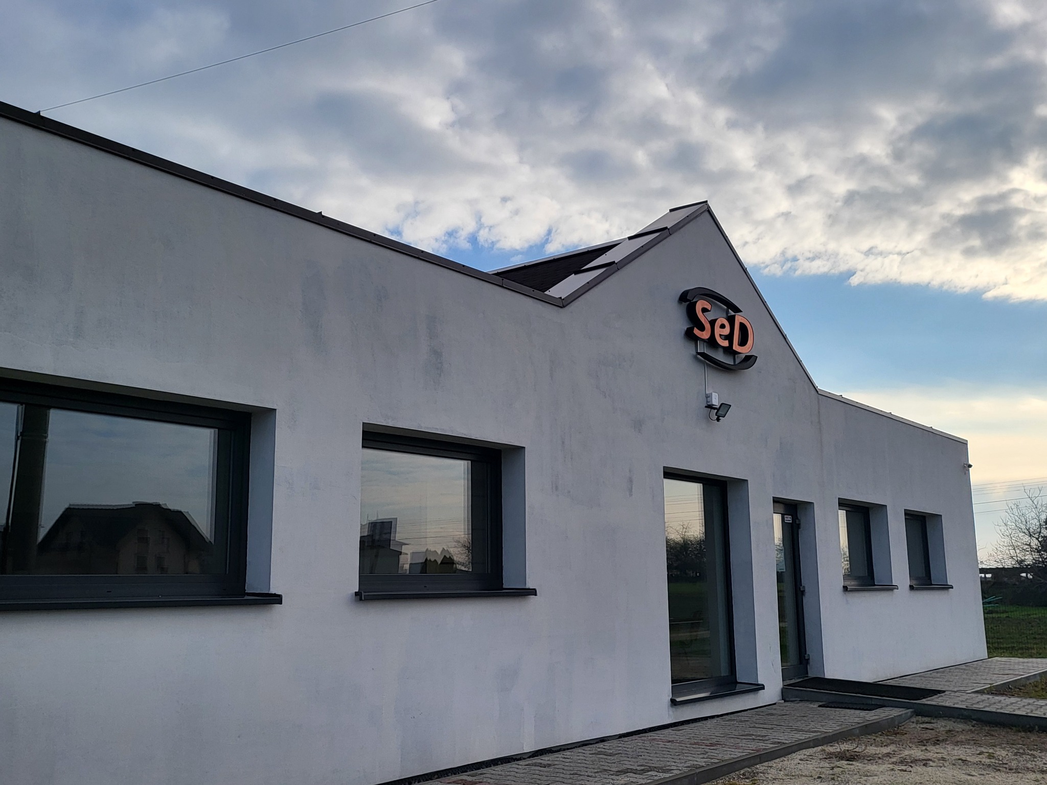 Biały budynek z ciemnymi oknami i logo 'SeD' na szczycie, widoczny z zewnątrz, na tle pochmurnego nieba. Ujęcie z poziomu chodnika.