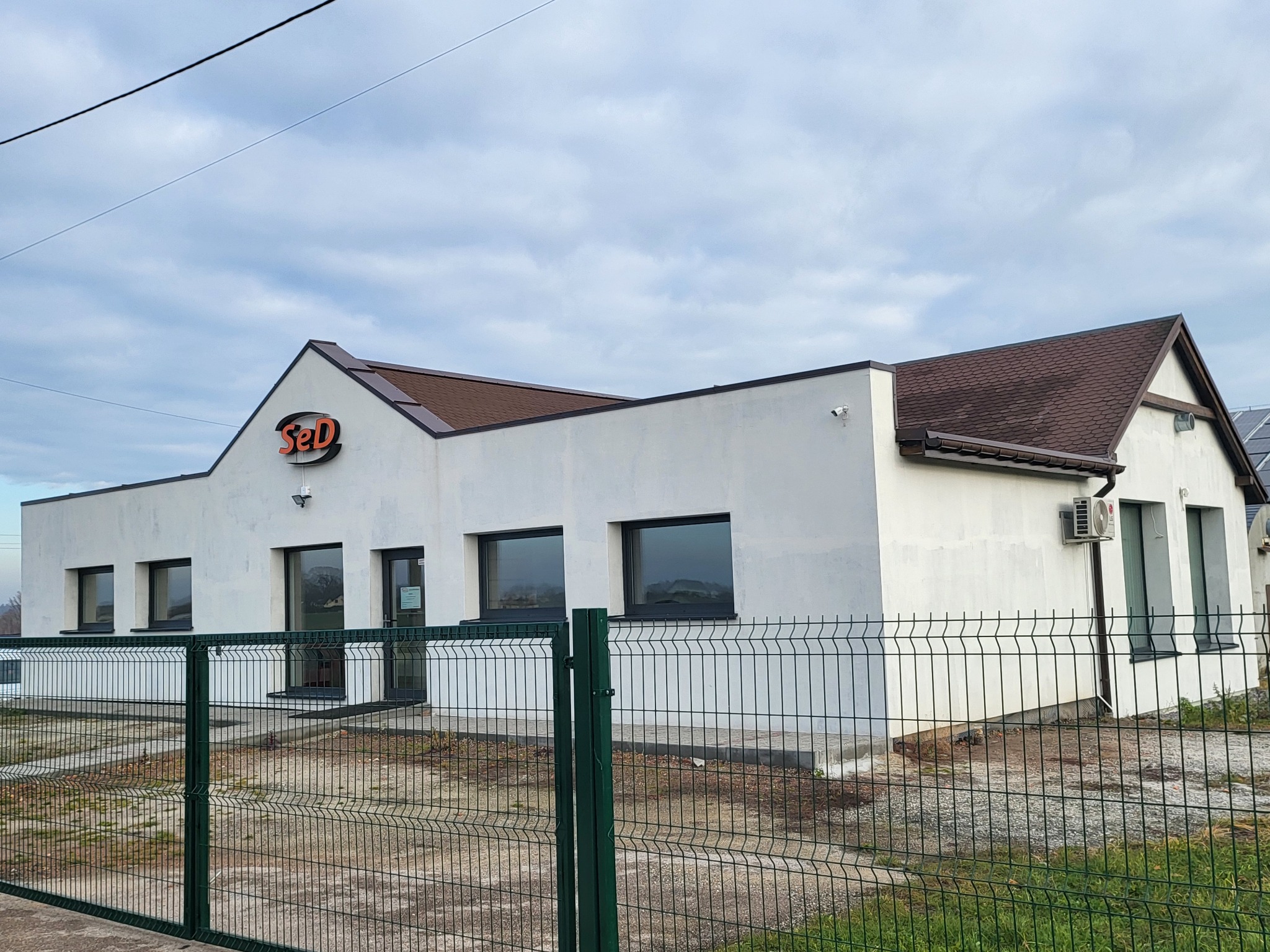 Biały budynek z brązowym dachem i logo 'SeD' nad wejściem, otoczony zielonym ogrodzeniem. Widoczny klimatyzator na ścianie i panele słoneczne w tle.