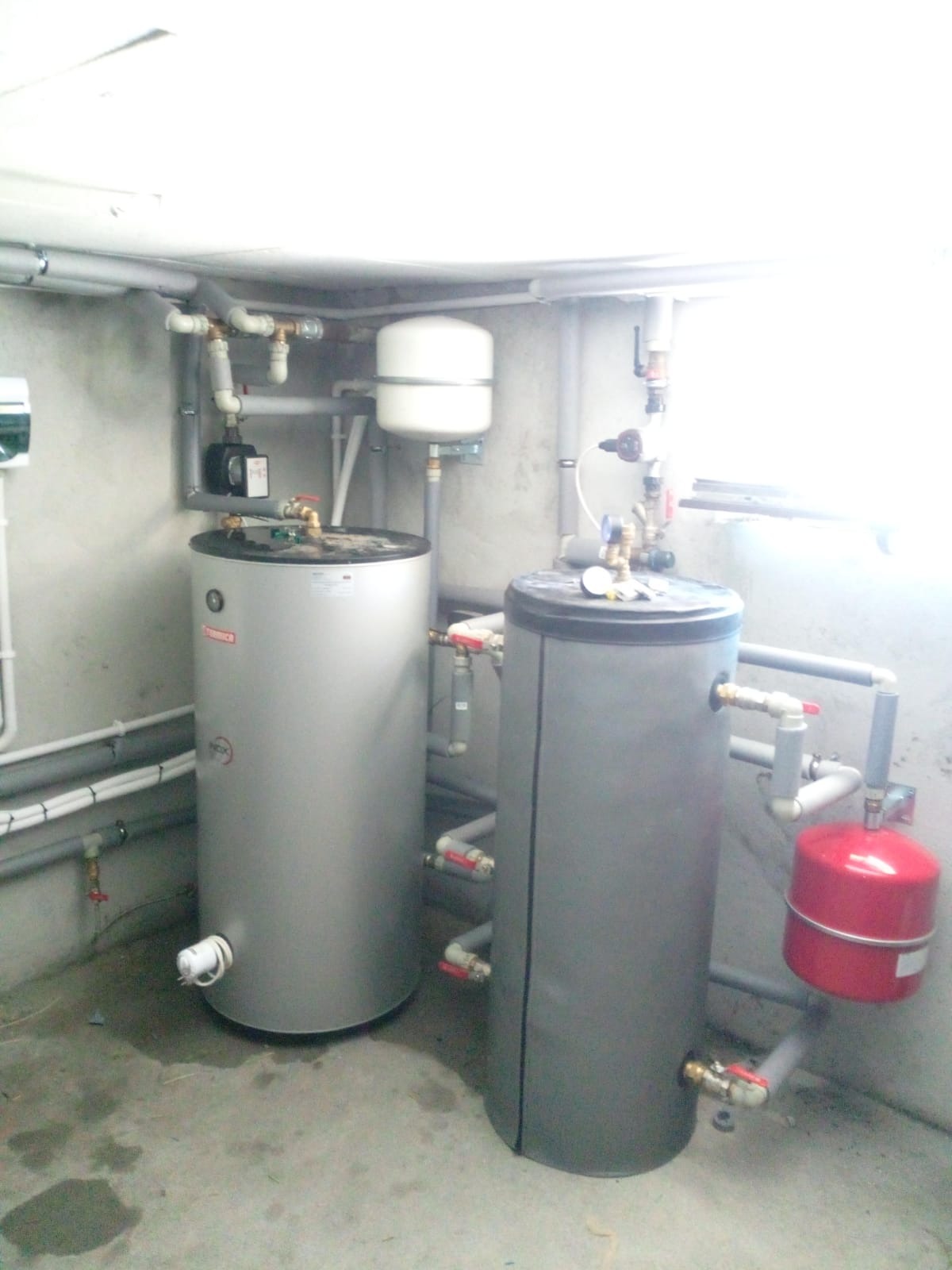 Dwa zbiorniki buforowe z instalacją hydrauliczną w kotłowni. Widoczne rury, zawory i osprzęt. Jeden zbiornik srebrny, drugi szary z czerwonym naczyniem wzbiorczym.