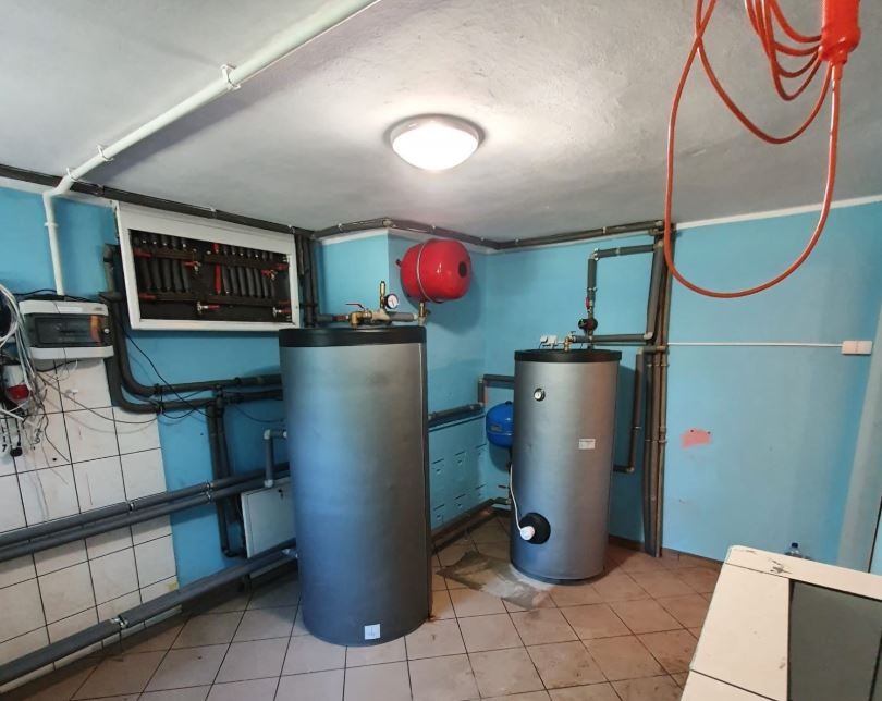 Dwa szare zbiorniki akumulacyjne w kotłowni z niebieskimi ścianami i brązowymi rurami. Widoczny panel sterowania i czerwony zbiornik wyrównawczy.