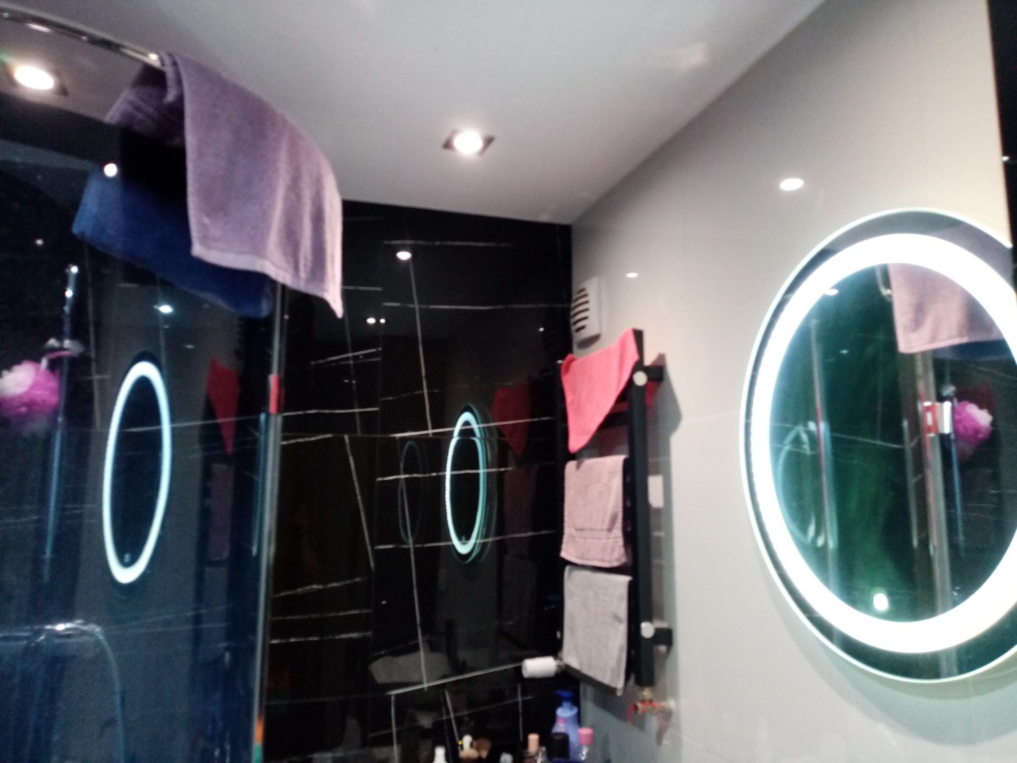 Nowoczesna łazienka z czarnymi błyszczącymi płytkami, okrągłym lustrem z podświetleniem LED i grzejnikiem drabinkowym z ręcznikami.