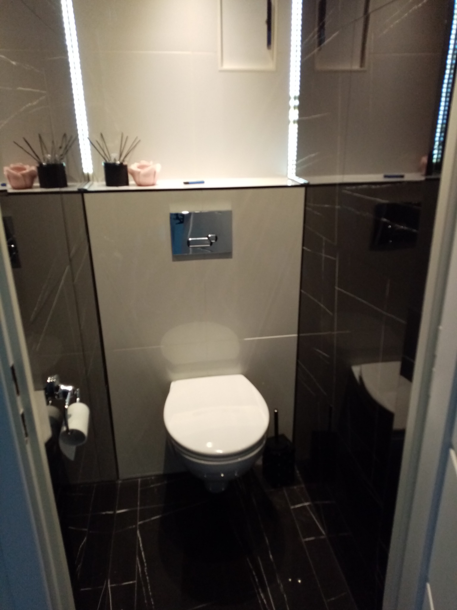 Nowoczesna łazienka w odcieniach szarości i bieli z wiszącą toaletą, czarną szczotką do WC i dekoracyjnymi dyfuzorami zapachowymi na półce.