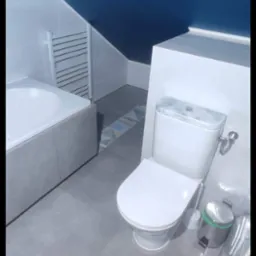 Nowoczesna łazienka z toaletą, wanną obudowaną szarą płytką i ciemnoniebieskim sufitem pod skosem, fragment dekoracyjnego pasa płytek na podłodze.