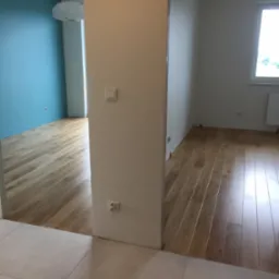 Jasne panele podłogowe ułożone w jodełkę w dwóch pomieszczeniach, widoczne listwy dystansowe, ściany pomalowane na biało i błękitnie, widok z korytarza wyłożonego płytkami.