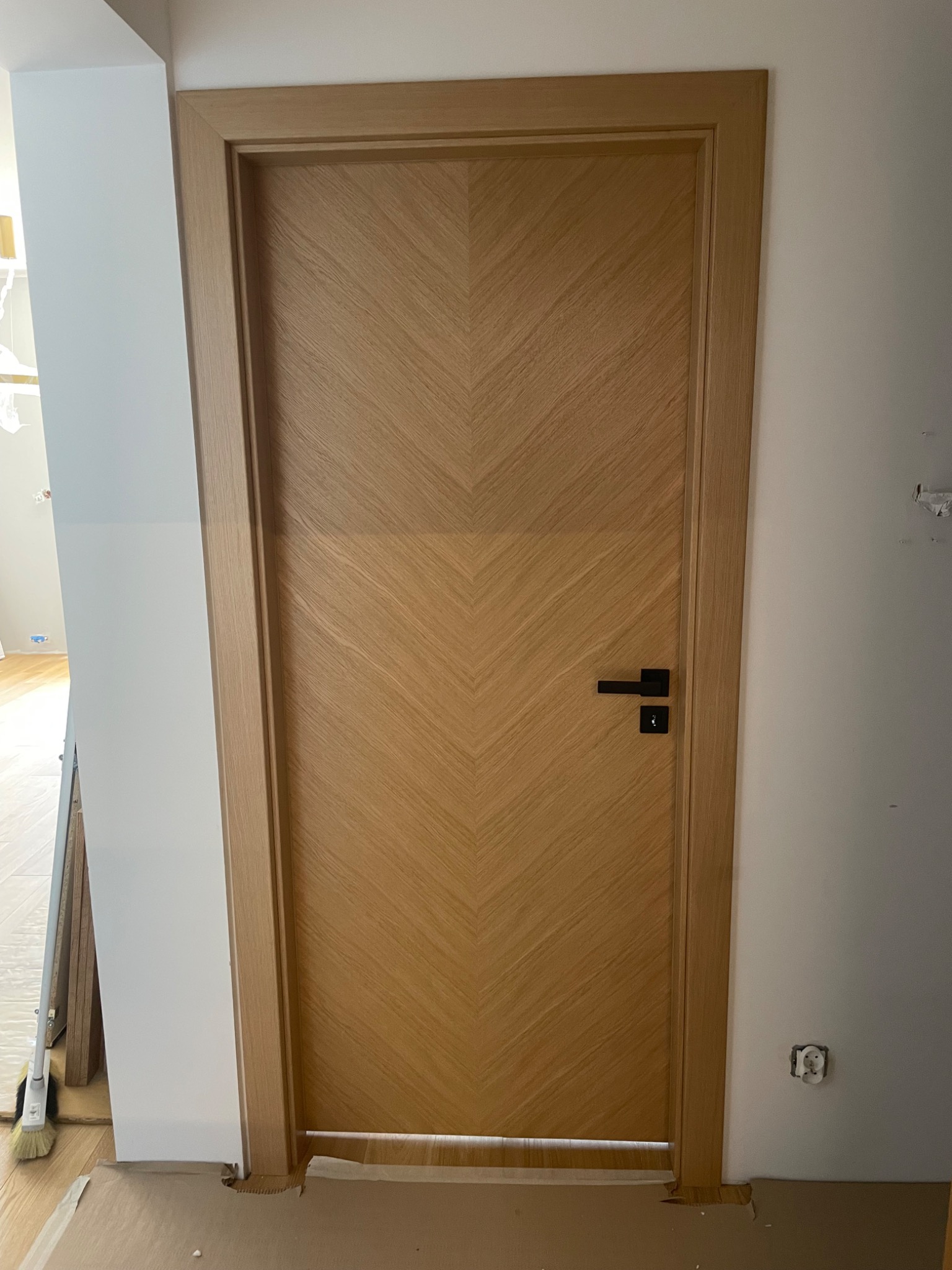 Zamknięte drzwi wewnętrzne z jasnego drewna z wzorem w jodełkę, czarna klamka, widoczne niedociągnięcia w wykończeniu ściany i podłogi.