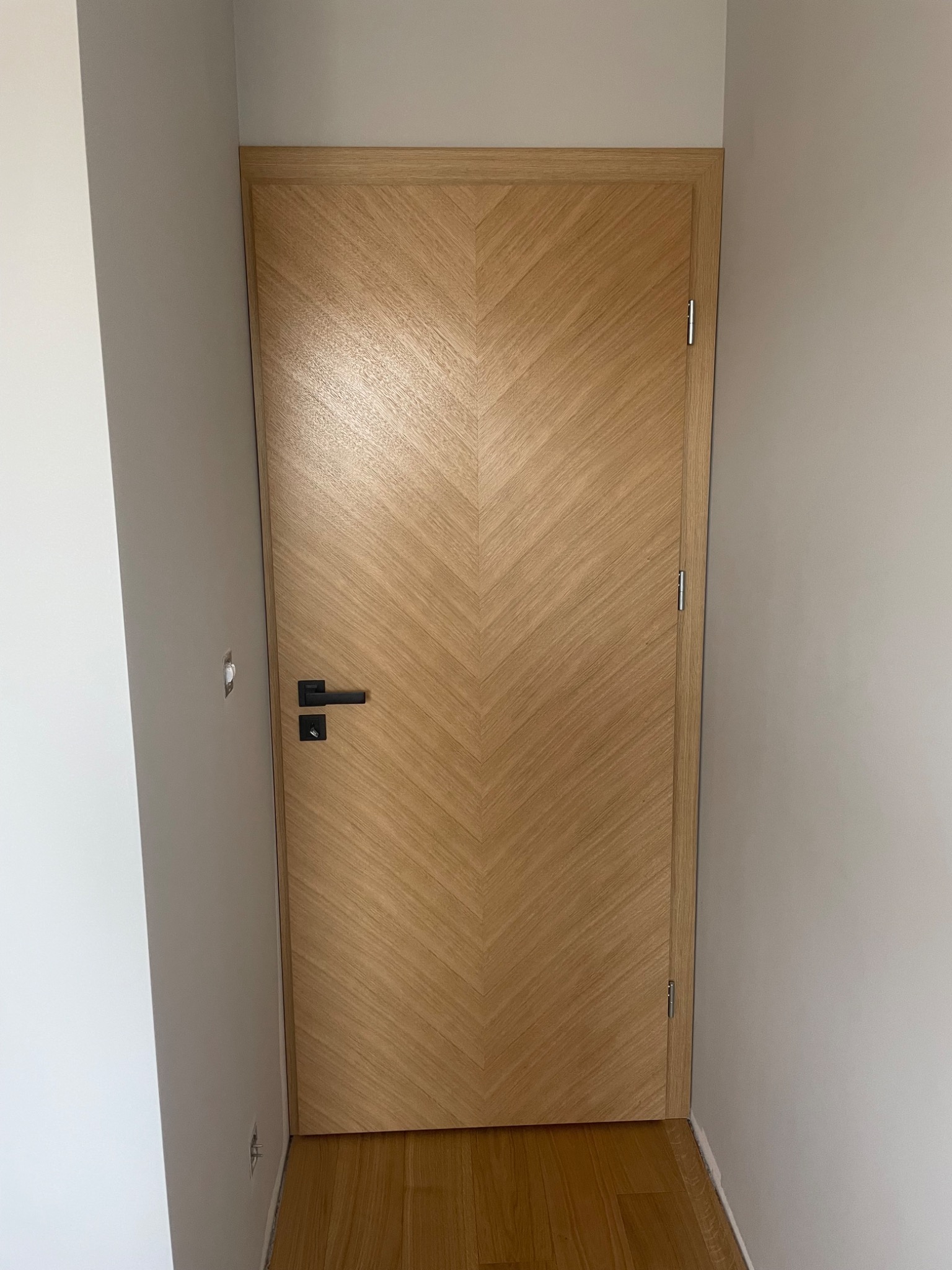 Zamknięte, drewniane drzwi z wzorem w jodełkę, czarna klamka, widoczne zawiasy, szara ściana po obu stronach.
