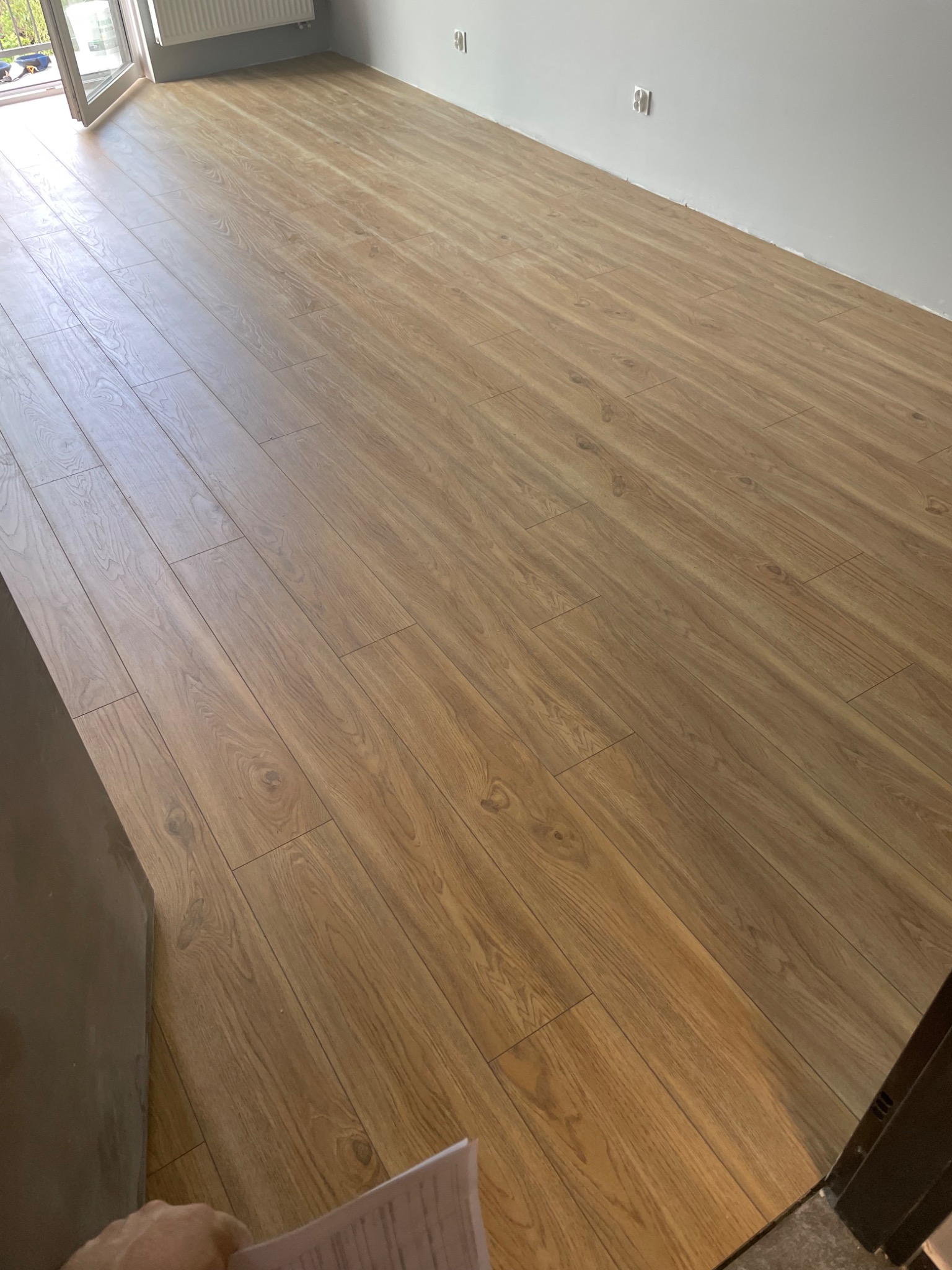 Ułożone panele podłogowe w jasnym odcieniu drewna, widoczne łączenia i struktura, fragment otwartego okna w tle.