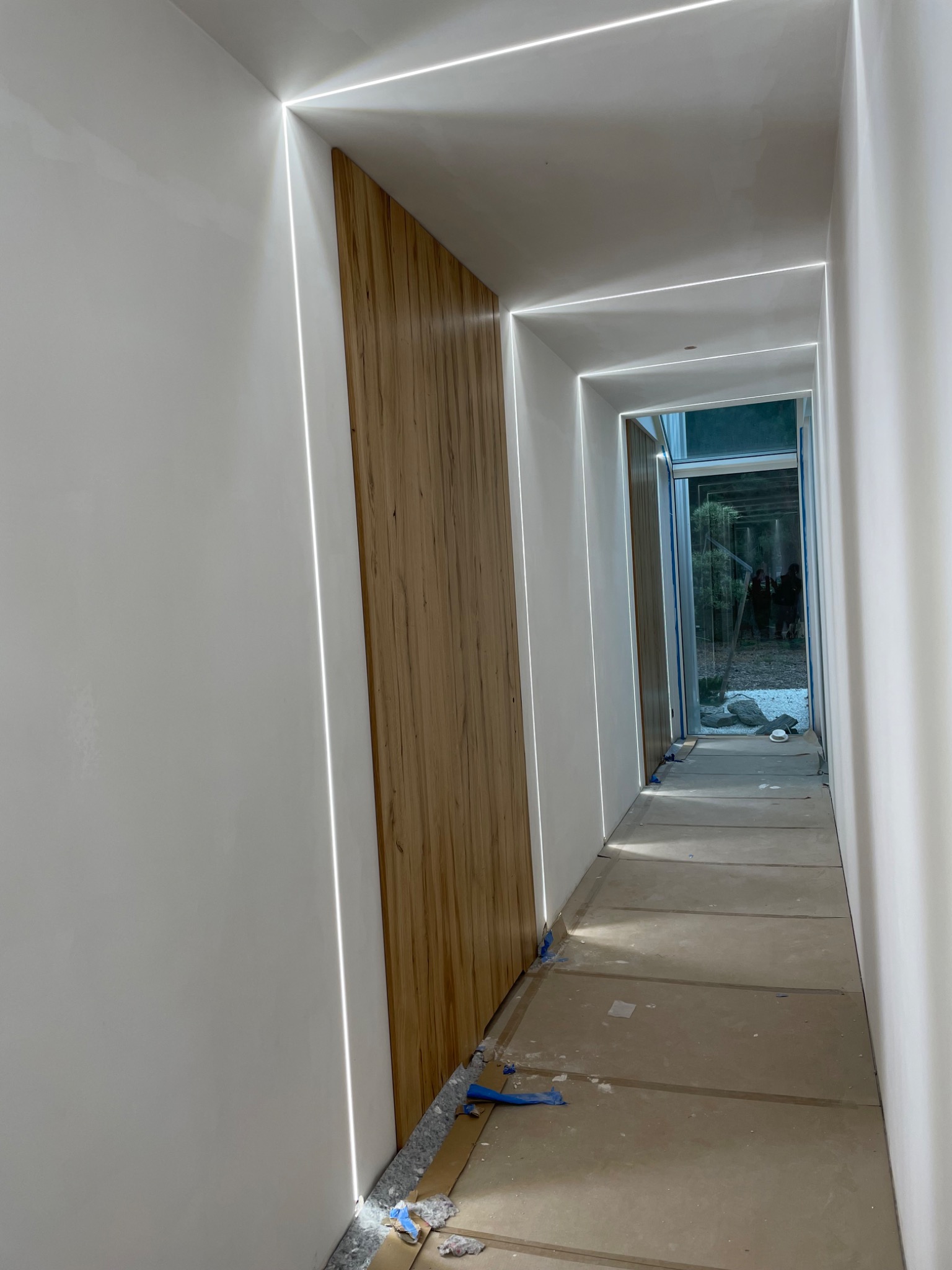 Korytarz w trakcie wykańczania z zamontowanymi panelami ściennymi z drewna i liniowym oświetleniem LED wzdłuż sufitu i ścian, zabezpieczona podłoga kartonem