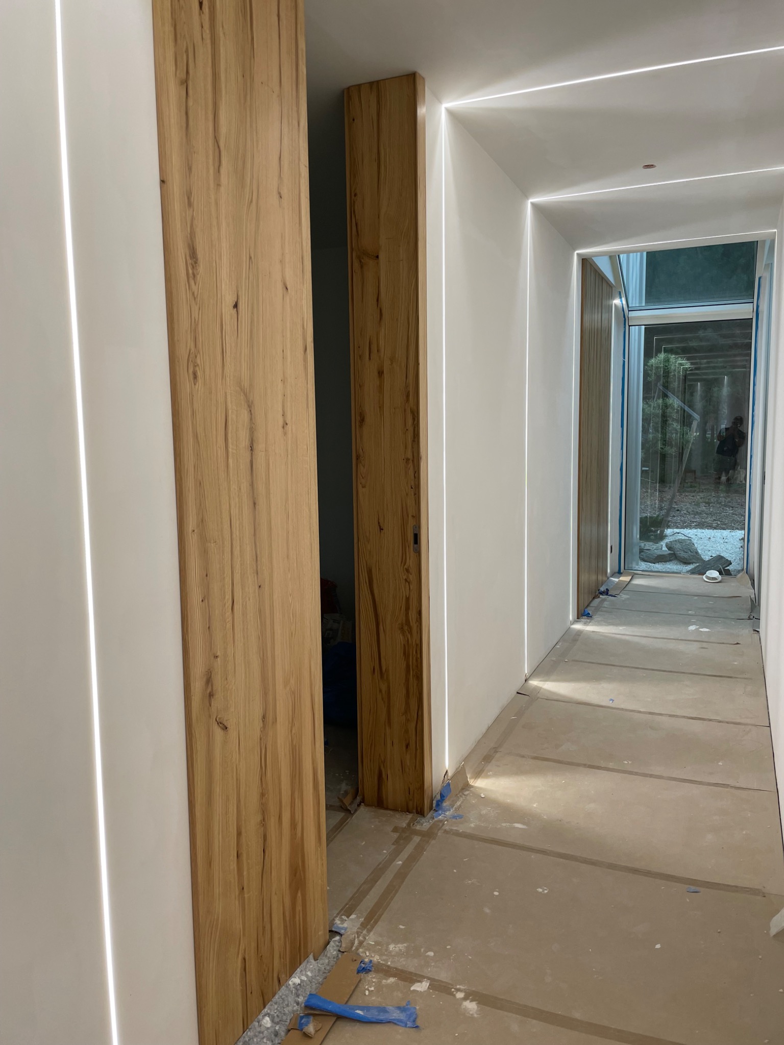 Nowoczesny korytarz z ukrytymi drzwiami z jasnego drewna i liniowym oświetleniem LED w ścianach i suficie, podłoga zabezpieczona kartonem podczas prac wykończeniowych.