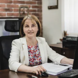 Kobieta w białym żakiecie uśmiecha się siedząc przy biurku z komputerem, dokumentami w segregatorze i długopisem, w tle ściana z cegły i meble biurowe.