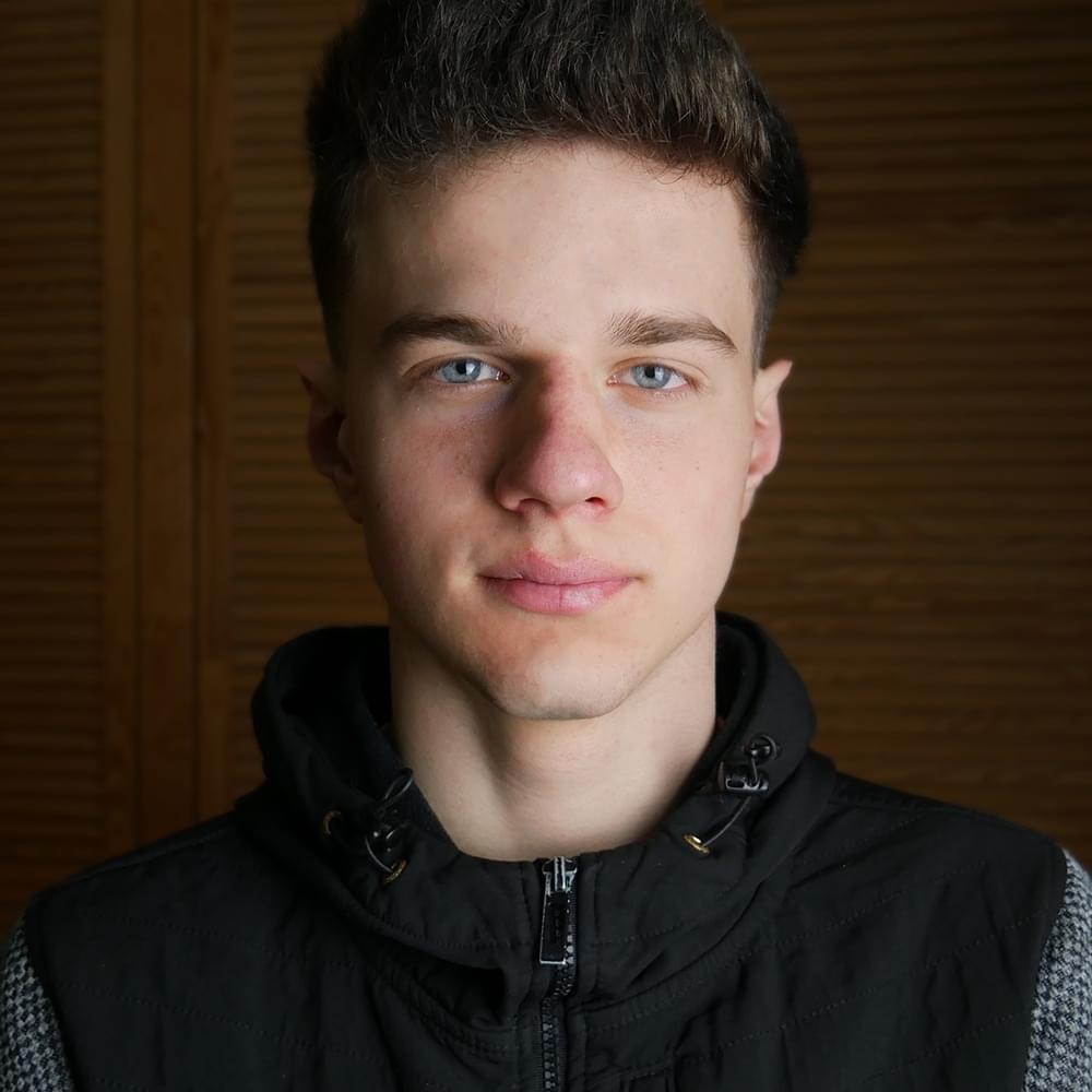 Portret młodego mężczyzny z niebieskimi oczami, ubrany w czarną kamizelkę na tle drewnianej ściany.