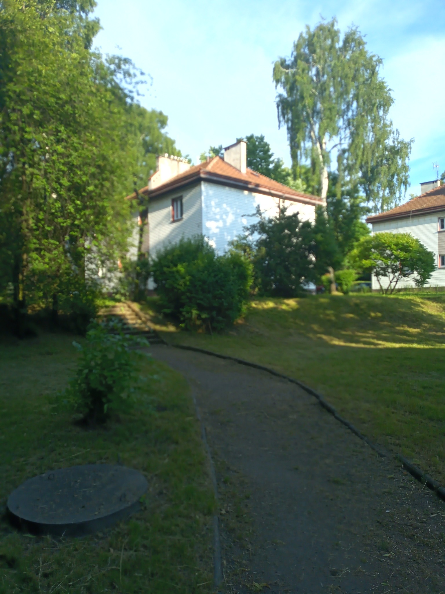 Ścieżka prowadząca przez trawnik w kierunku budynku mieszkalnego otoczonego zielenią, z widocznym włazem kanalizacyjnym na pierwszym planie.