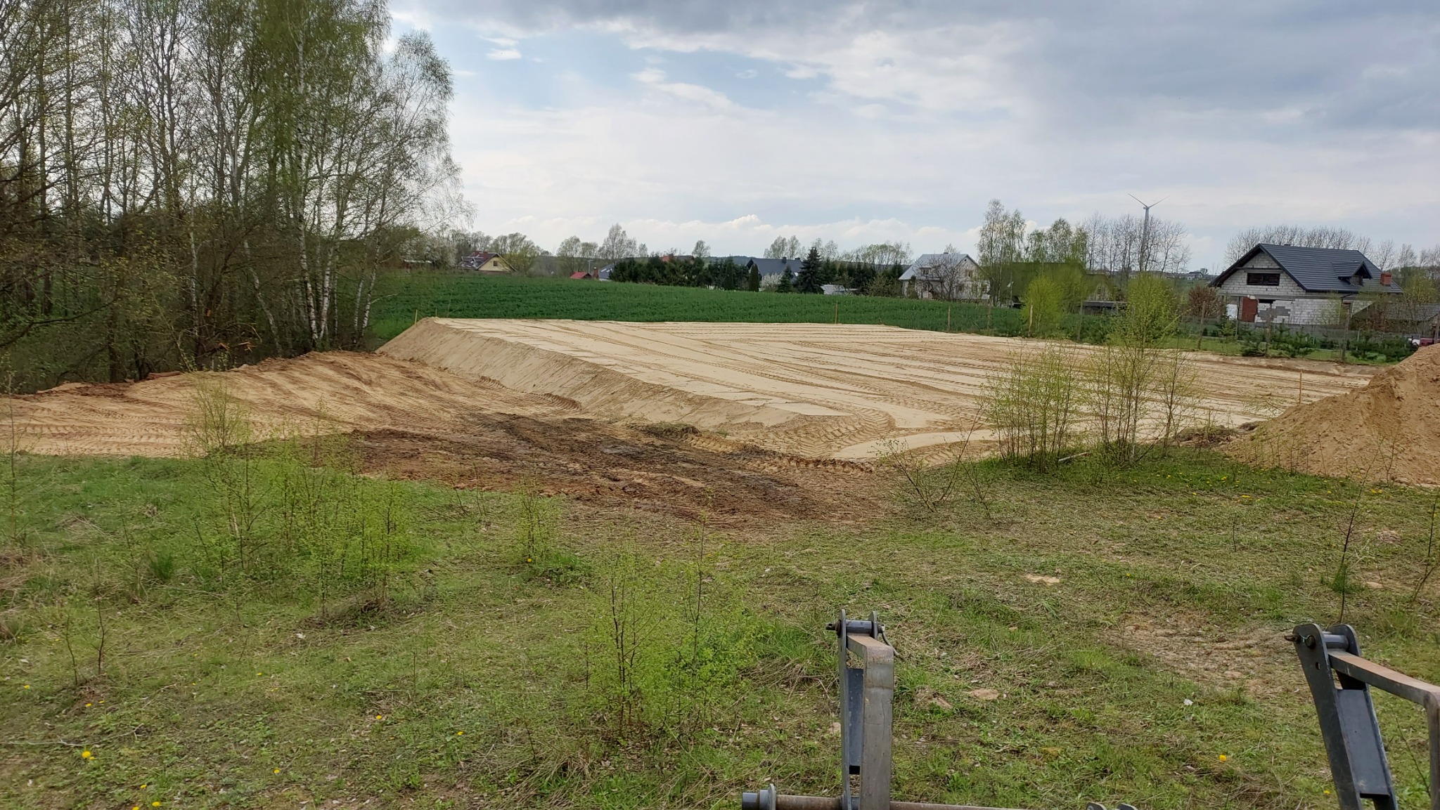 Przygotowany teren pod budowę z widocznymi śladami opon, usypane kopce piasku, w tle zabudowa jednorodzinna i drzewa.